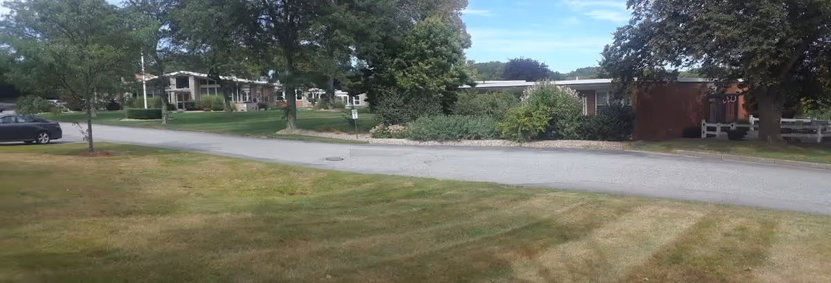 Grassy lawn and a paved road lined with trees and low residential buildings on a senior living campus.