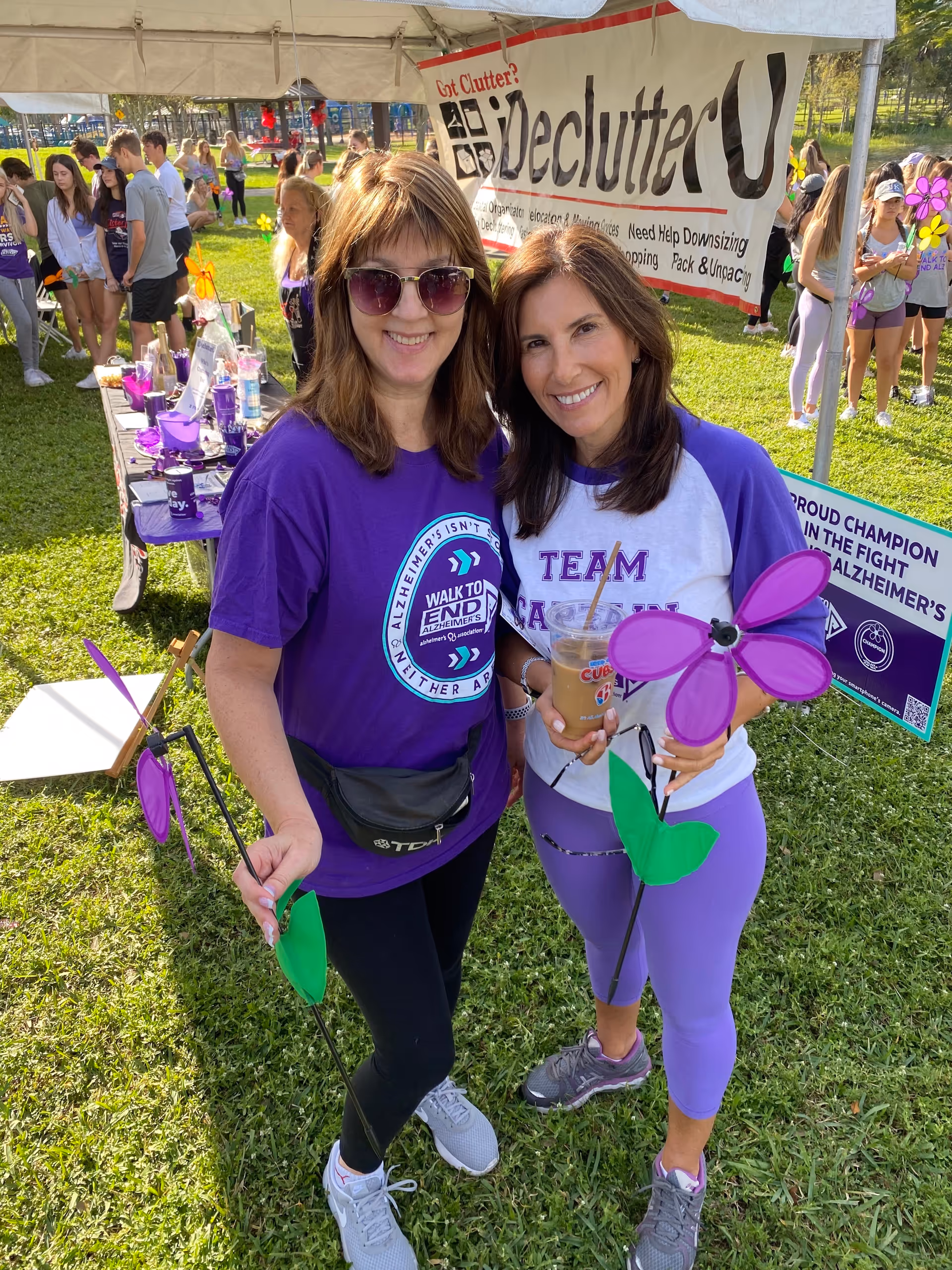 Two women in purple shirts pose outdoors at a park fundraiser under a banner.