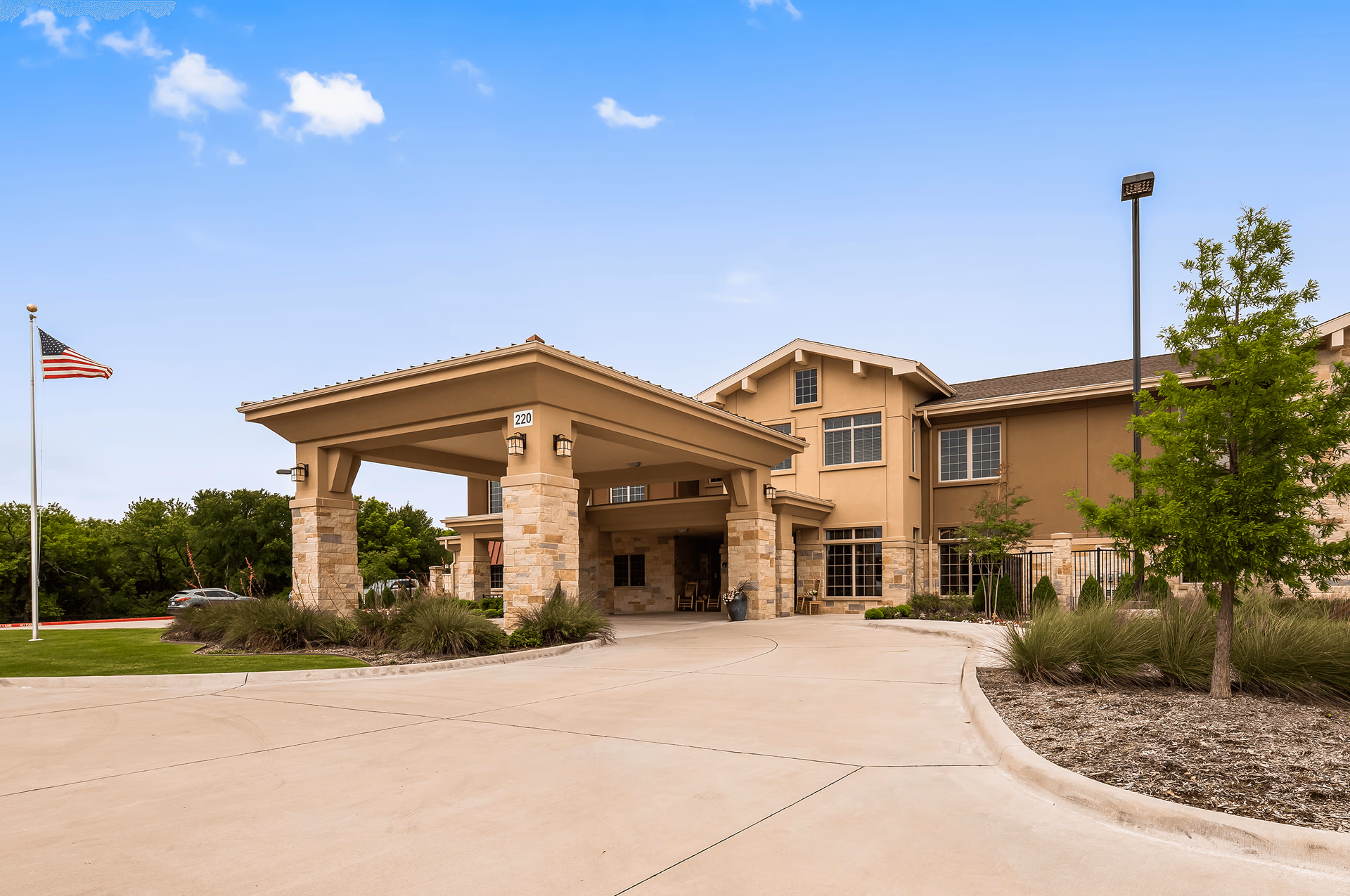 Exterior view of Legend of McKinney facility showing the entrance with a covered driveway supported by stone pillars, a flagpole with an American flag, and surrounding greenery under a blue sky with a few clouds.