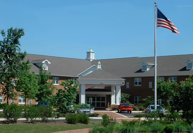Brick senior living facility exterior with a white entrance, flagpole, and several parked cars.