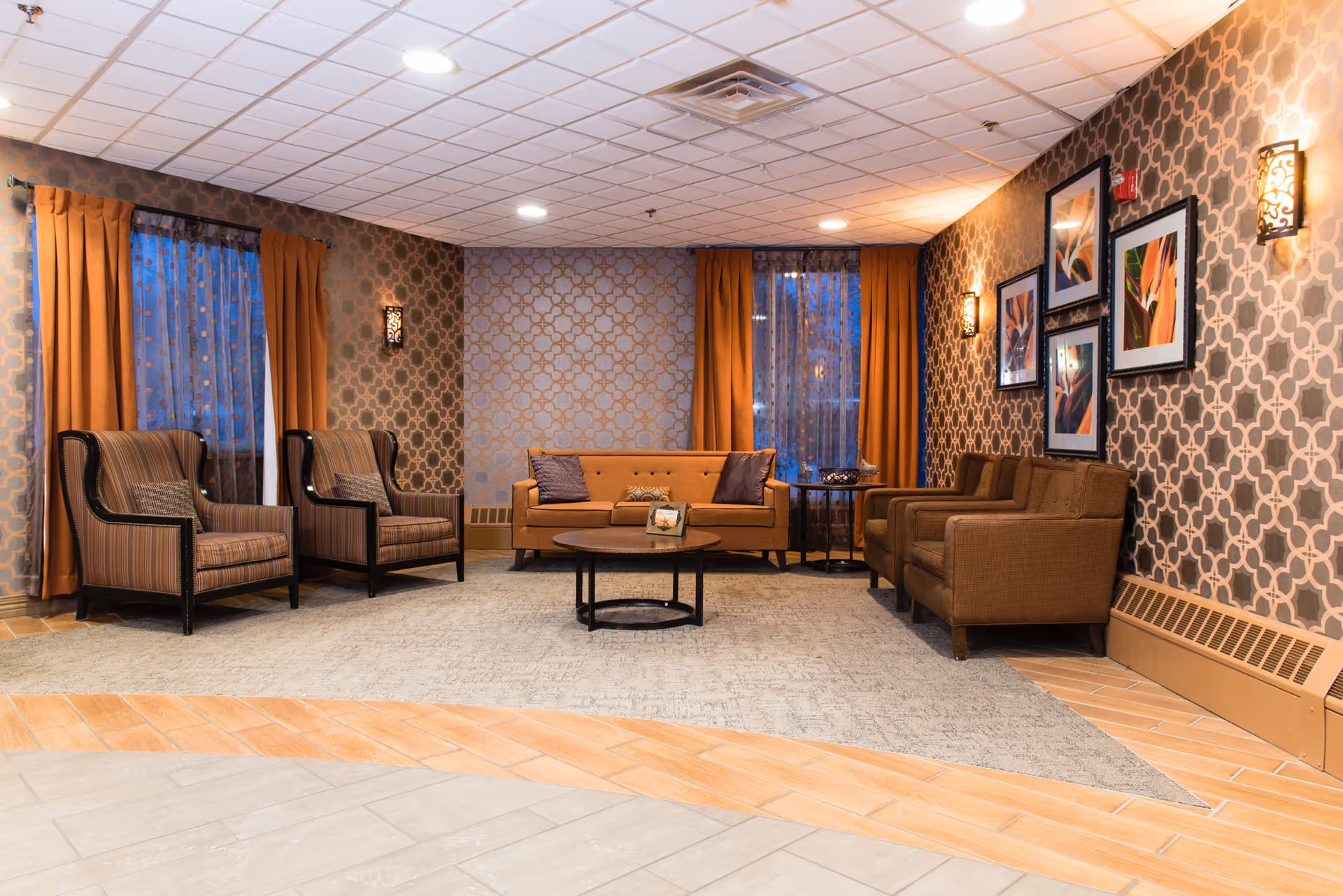 A warmly lit senior living lounge with brown seating, patterned wallpaper, and orange curtains.