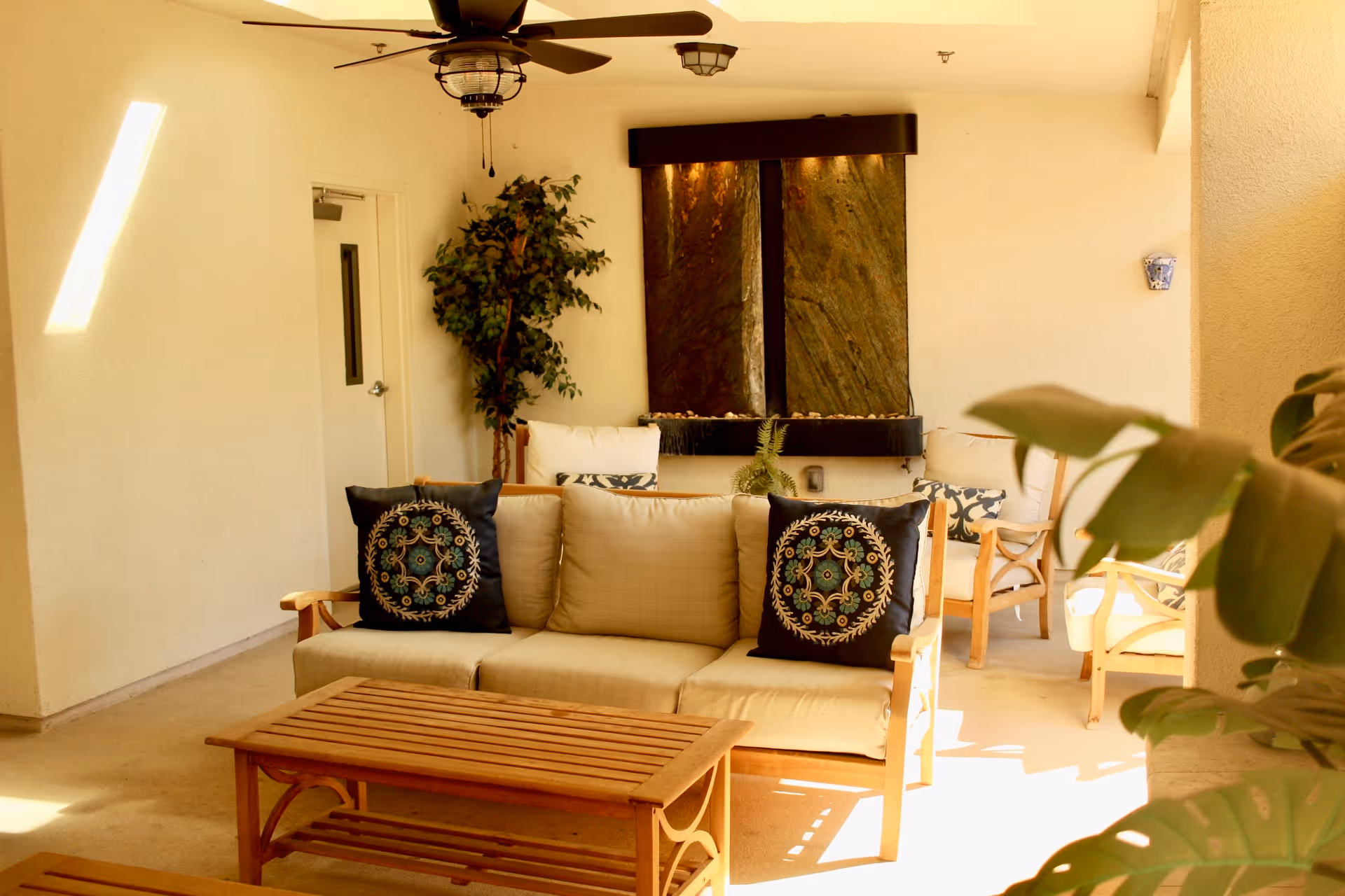 A cozy indoor seating area with a beige cushioned sofa adorned with two decorative pillows featuring intricate patterns. In front of the sofa is a wooden coffee table. Behind the sofa, there is a water feature with two vertical stone panels and a small plant. To the left, a potted artificial tree stands near a white door. Ceiling fans with lights hang from the ceiling, and sunlight streams in from the right side, illuminating the space.