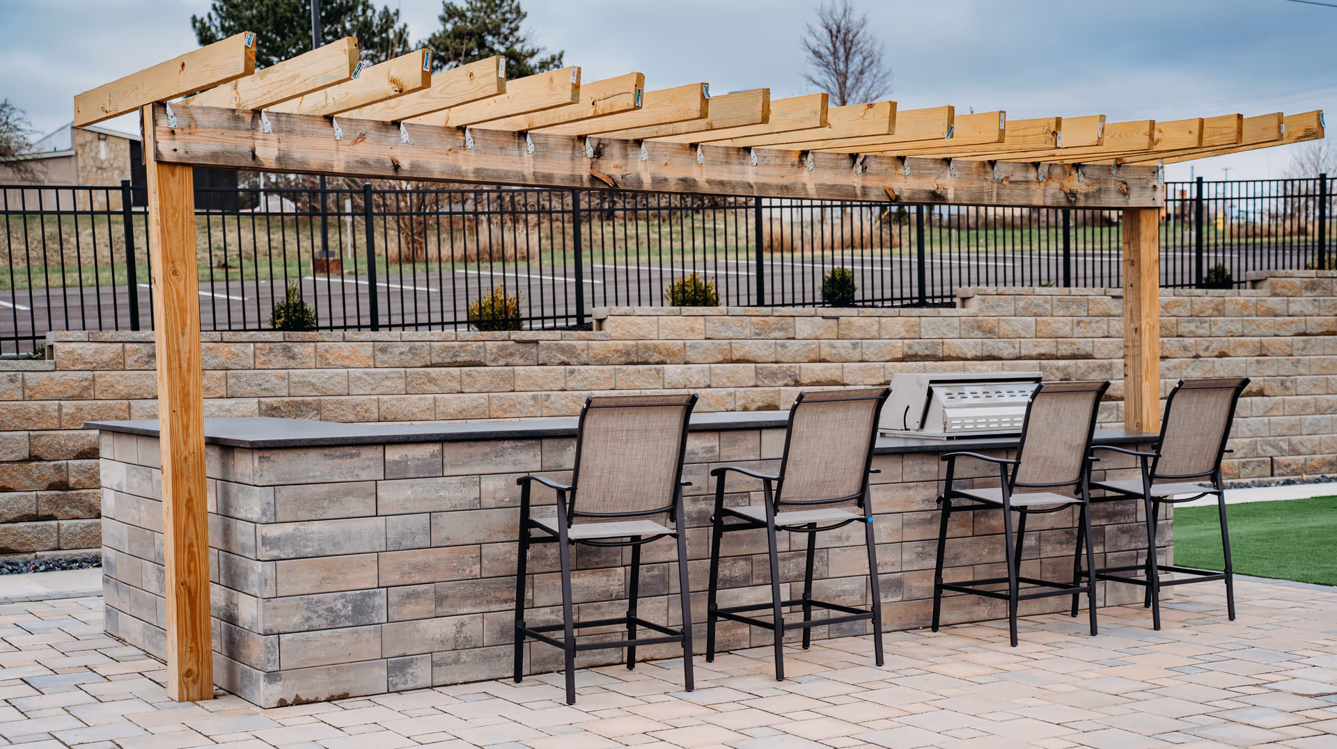 Outdoor patio bar with a wooden pergola, five high chairs, and a built-in grill in front of a stone retaining wall.