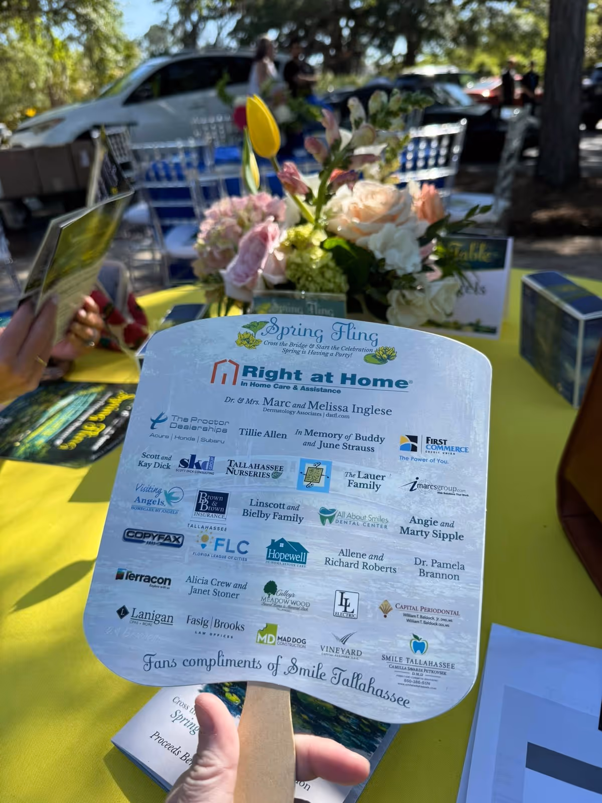 Close-up of a hand holding a curved glossy event sign on a yellow outdoor table.