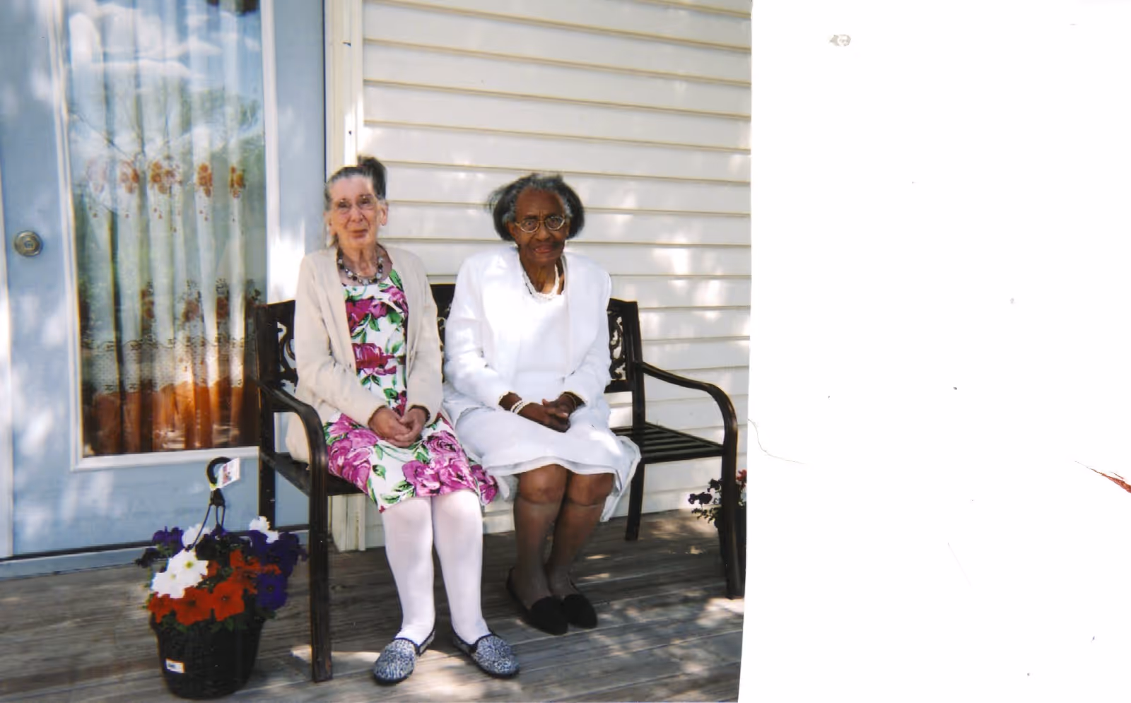 Two elderly women sitting on a black metal bench on a wooden porch outside a house with white siding. One woman is wearing a floral dress with a beige cardigan and white tights, and the other woman is dressed in a white outfit with black shoes. There is a basket of colorful flowers on the porch floor next to the bench.