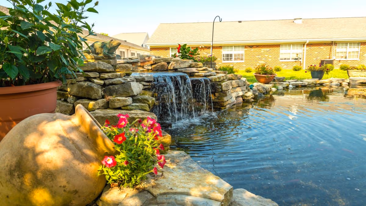 A landscaped outdoor pond with a stone waterfall, potted flowers, and a brick building in the background.
