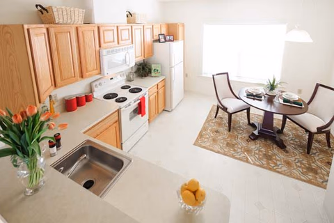 Bright kitchen and dining area with wooden cabinets, white appliances including a stove, microwave, and refrigerator. A round dining table with two chairs is set on a patterned rug near a large window letting in natural light. The kitchen counter has a sink, a vase with orange tulips, and a bowl of lemons.