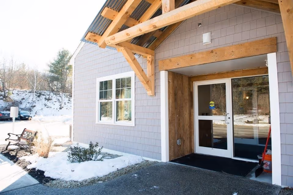Entrance to a building with a wooden overhang and glass double doors. There is snow on the ground and a bench near the entrance. Trees and parked cars are visible in the background.