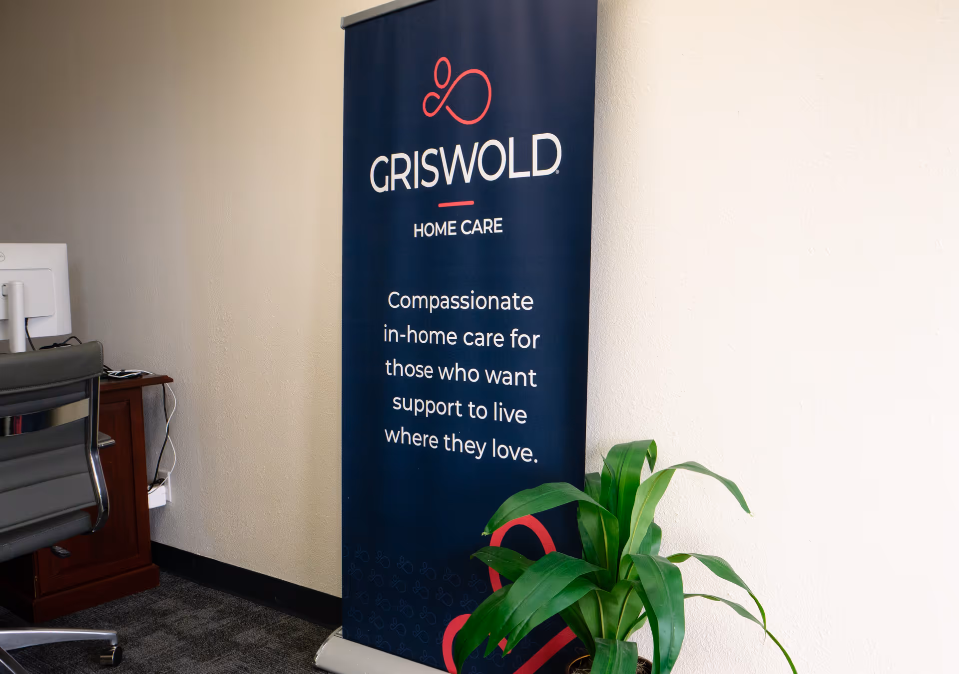 Tall Griswold Home Care banner beside desk, plant, and office chair in an interior.