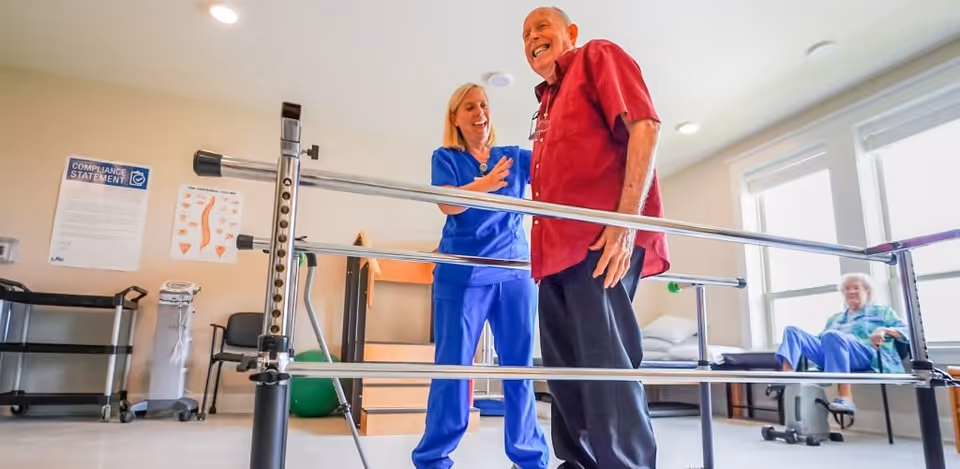 A healthcare professional in blue scrubs assists an elderly man in a red shirt as he practices walking between parallel bars in a rehabilitation or physical therapy room. In the background, an elderly woman sits on a chair near large windows, and various medical and exercise equipment are visible.