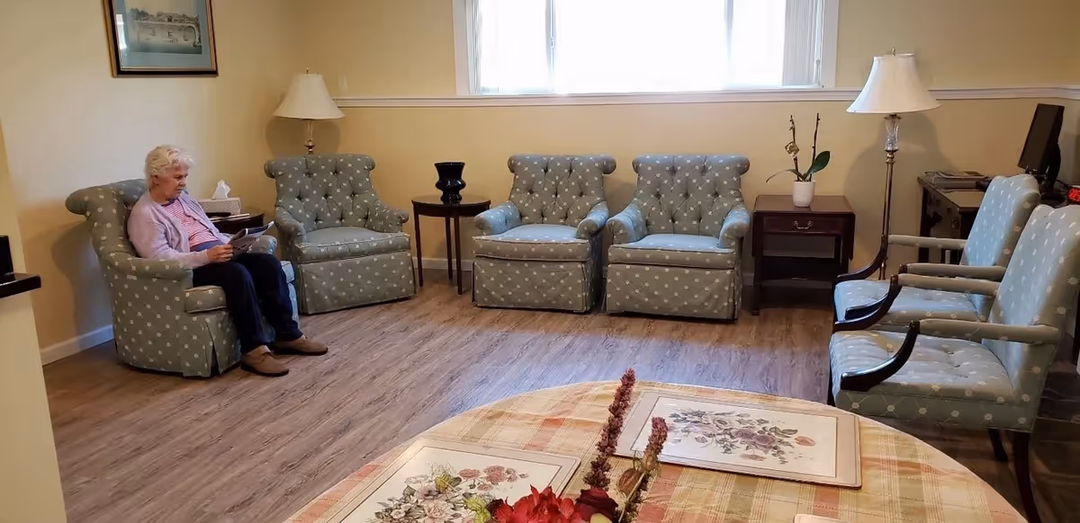Senior living room with blue patterned armchairs and a resident reading beside a table.