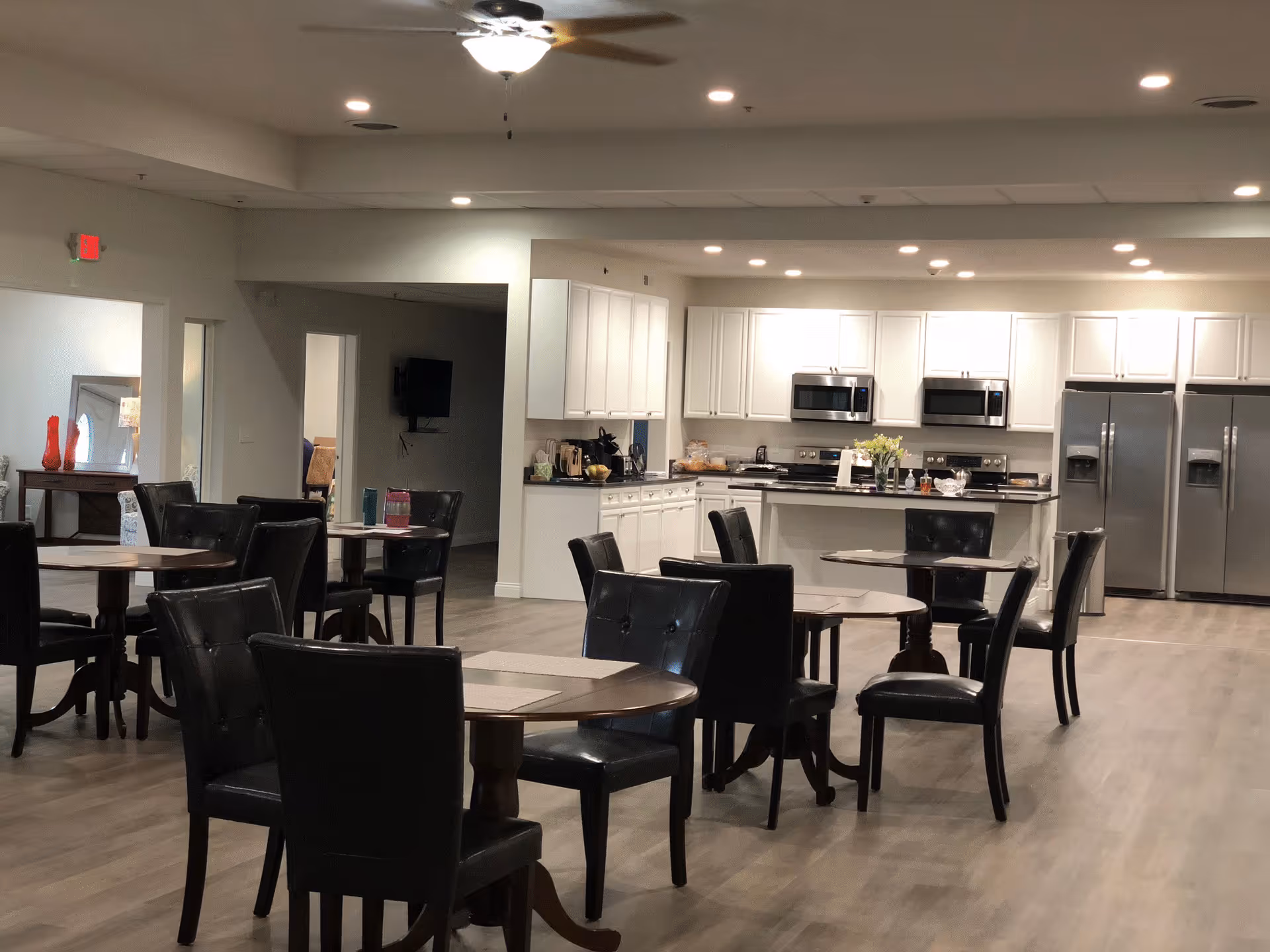 A spacious dining area with several round tables and black cushioned chairs. In the background, there is a modern kitchen with white cabinets, stainless steel appliances including two refrigerators, two microwaves, and a stove. The room has light-colored wooden flooring and recessed ceiling lights.