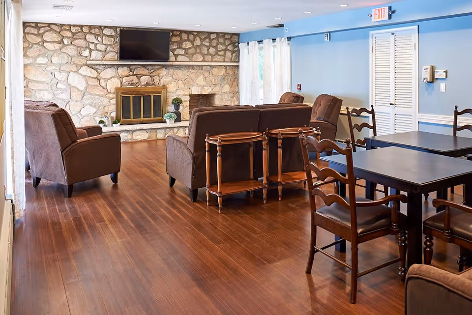 A cozy living room area with brown upholstered armchairs and sofas arranged around a stone fireplace with a mounted flat-screen TV above it. There are wooden side tables next to the sofas and dark wooden dining tables with chairs on the right side. The room has wooden flooring, light blue walls, and white curtains on the windows.