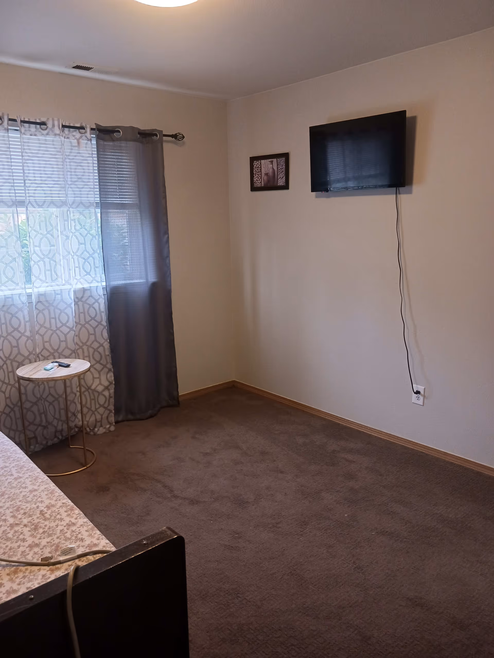 Sparsely furnished bedroom with a wall-mounted TV, patterned curtains, a small round side table, and carpeted floor.