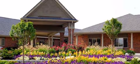 Brick senior living facility entrance with colorful flower beds and small trees.