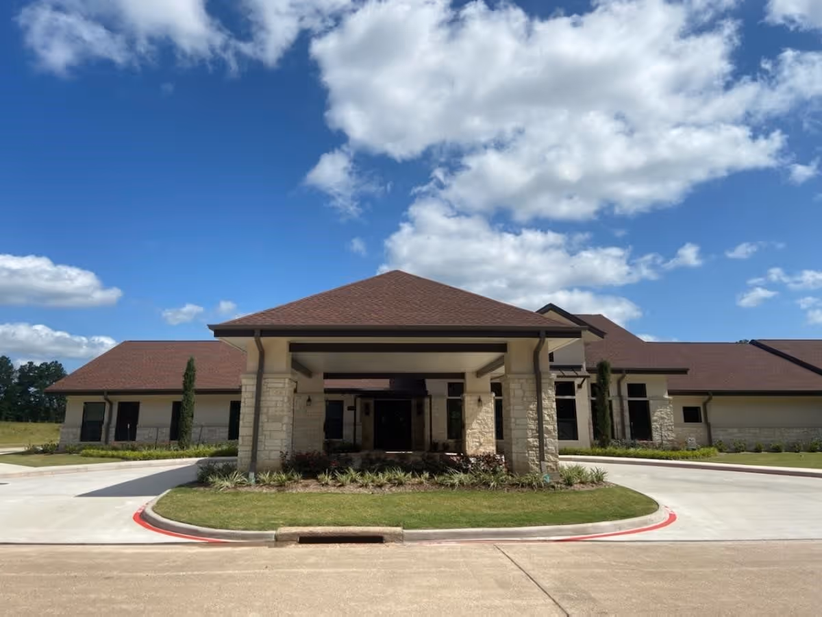 Front exterior view of The Bradford Memory Care and Assisted Living facility showing a single-story building with a covered entrance, stone pillars, and a brown roof under a partly cloudy blue sky.