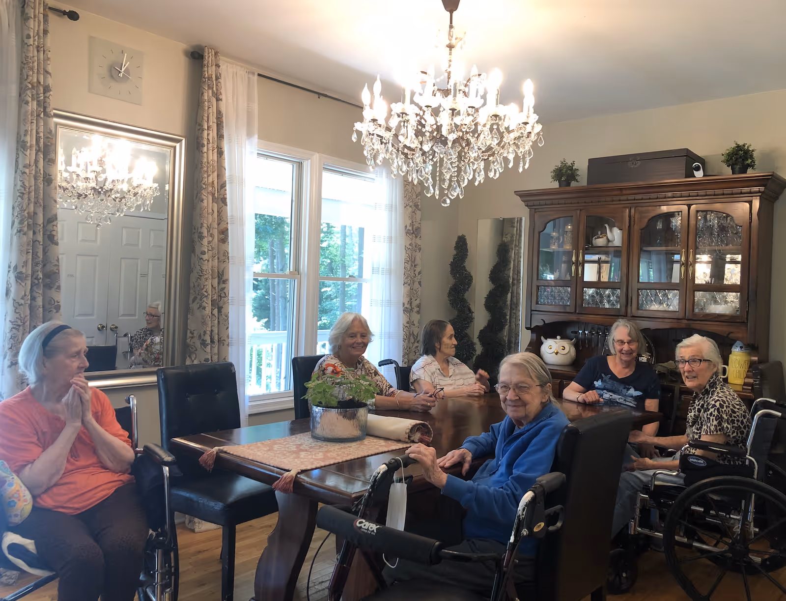 A group of six elderly women sitting around a wooden dining table in a well-lit room with large windows and floral curtains. The room features a crystal chandelier, a large mirror, and a wooden cabinet with glass doors displaying dishes and decorative items. Some women are seated in wheelchairs and appear to be engaged in conversation.