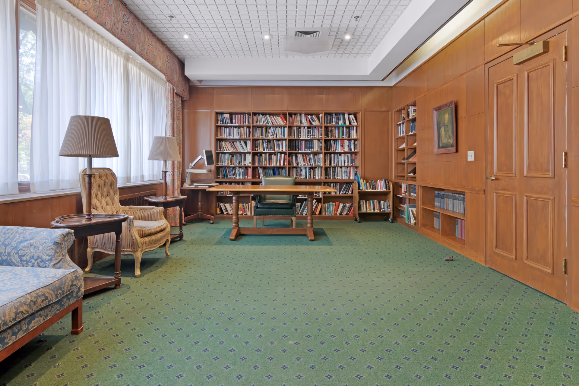 A quiet library room with green patterned carpet, wooden paneled walls, and a large bookshelf filled with books. There is a wooden table with a green chair in front of the bookshelf, two upholstered armchairs with side tables and lamps near a large window with sheer white curtains, and a blue patterned sofa on the left side.