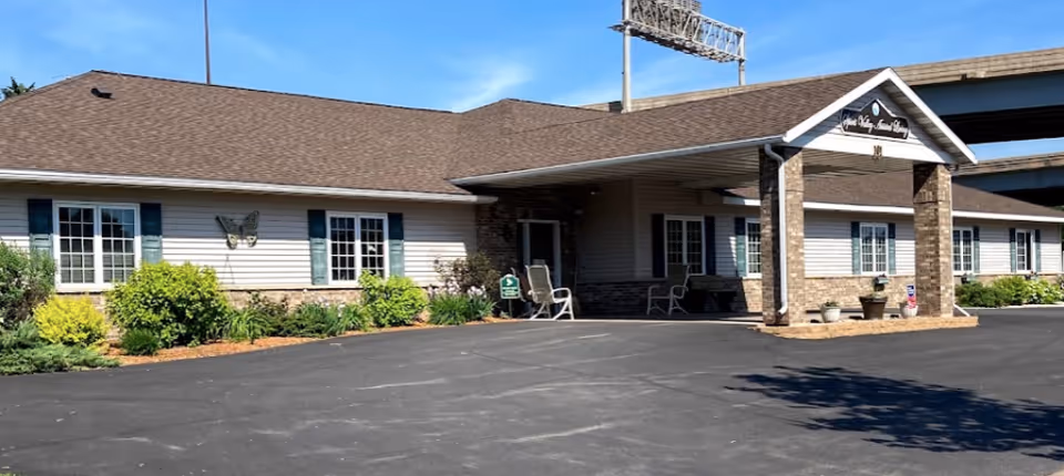Single-story senior living building exterior with a covered entrance, chairs, windows, and landscaping under a clear blue sky.