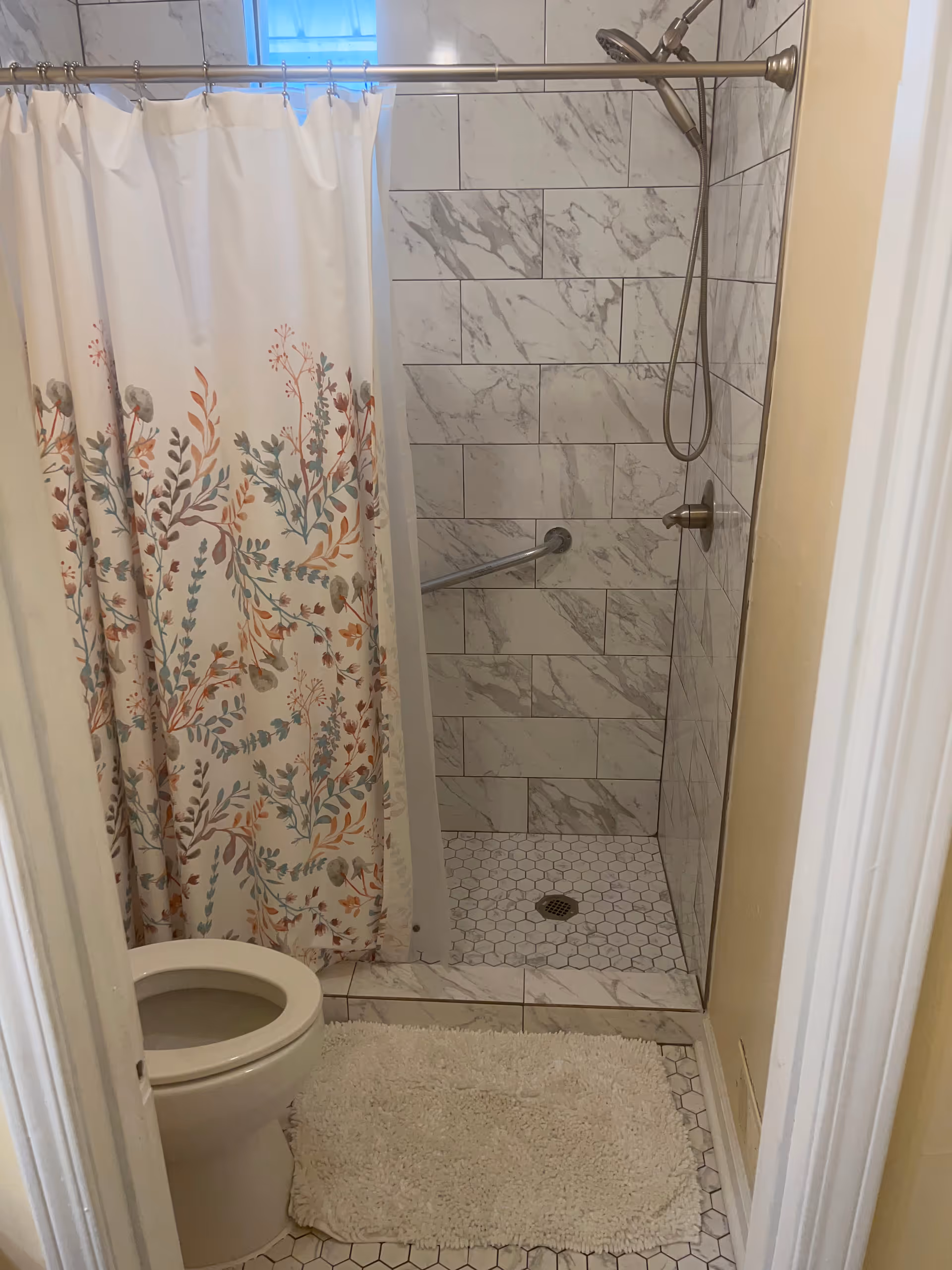 A small bathroom featuring a toilet with the lid open, a white and floral patterned shower curtain, a tiled shower area with marble-like tiles on the walls and hexagonal tiles on the floor, a silver showerhead, and a white bath mat on the floor.