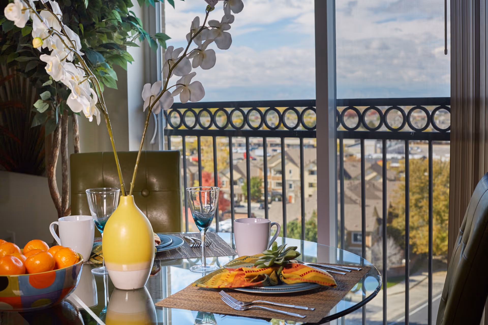 Glass dining table with colorful settings, vase, fruit bowl, and balcony view.
