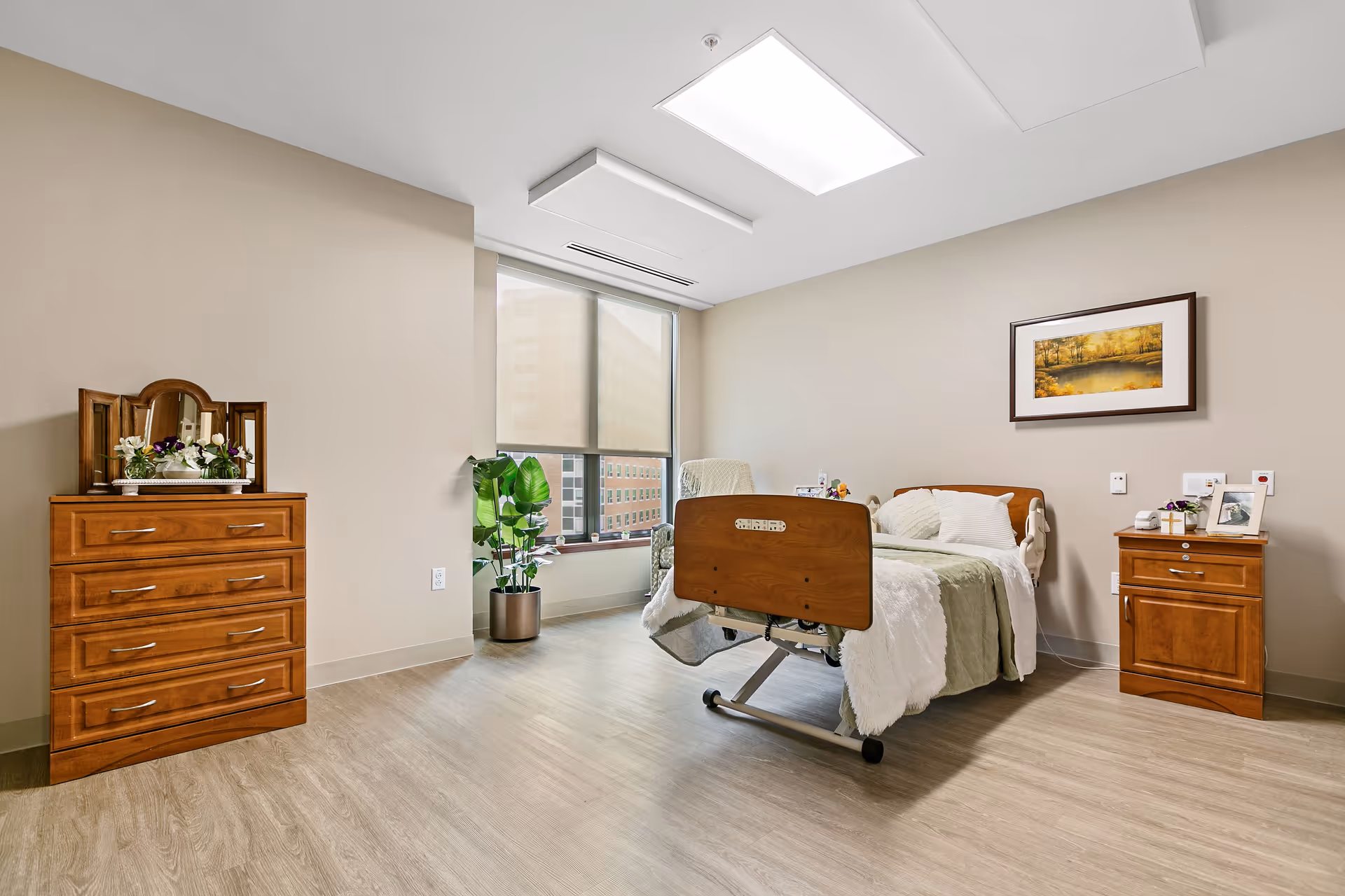 A furnished senior living bedroom with a hospital-style bed, dressers, and a window.