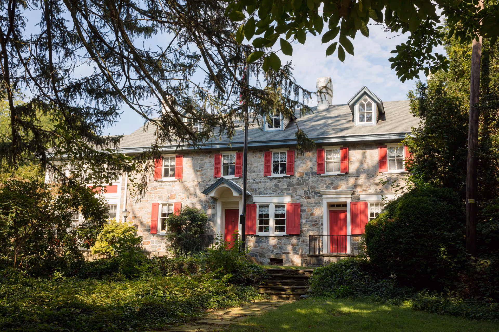 Stone two-story building with red shutters and lush greenery surrounding the front entrance.