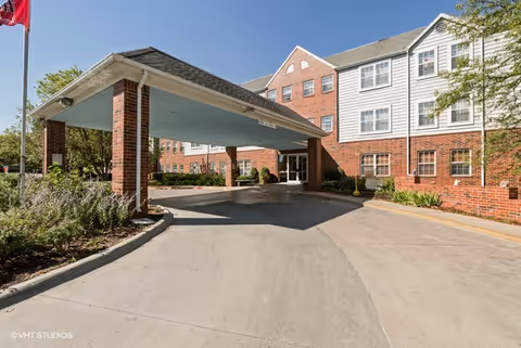 Entrance area of a senior living facility with a covered drop-off zone supported by brick columns. The building is made of red brick with white-trimmed windows and a gabled roof. There are some shrubs and trees around the driveway under a clear blue sky.