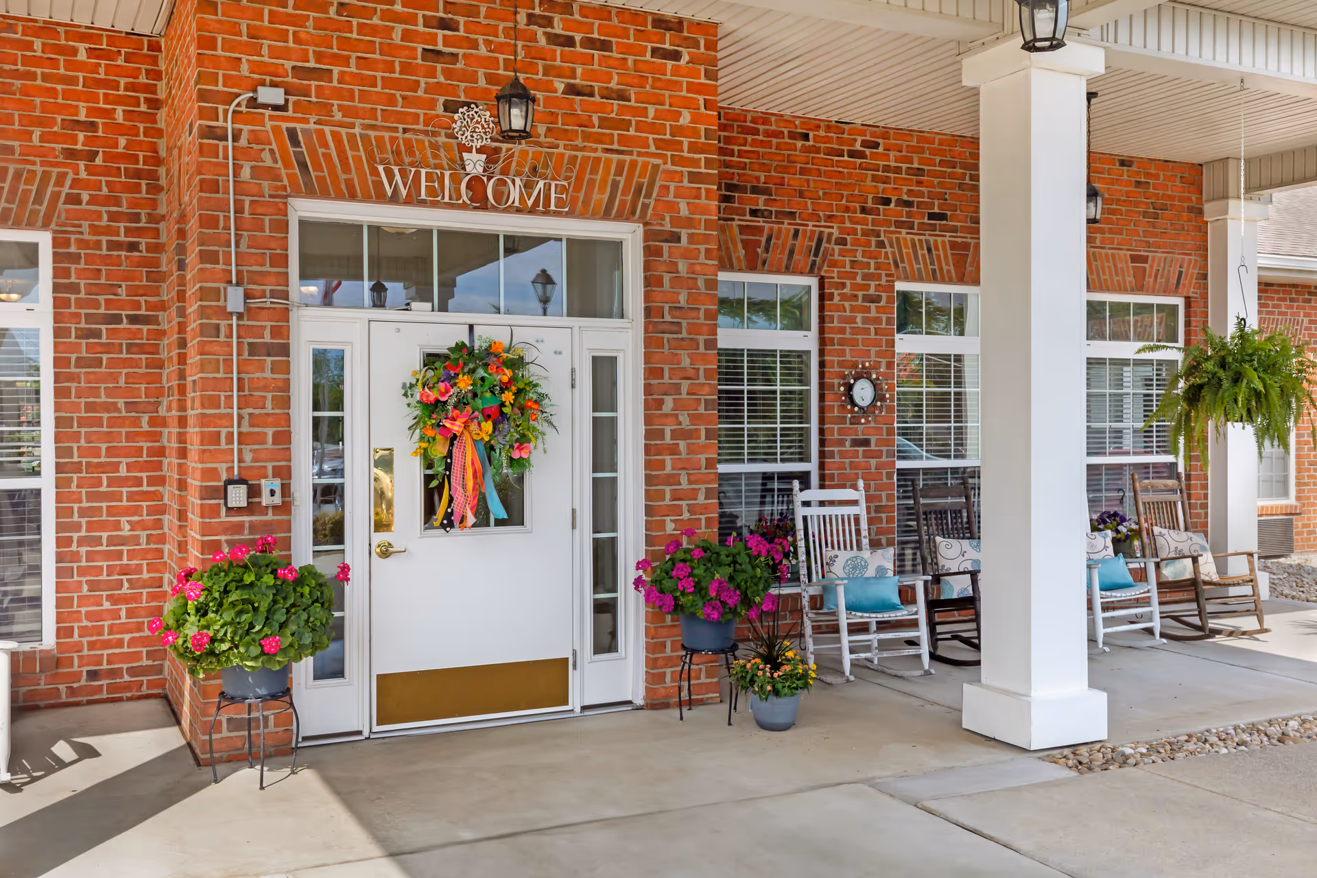 Brick building entrance with white door, welcome sign, porch seating, and potted flowers.