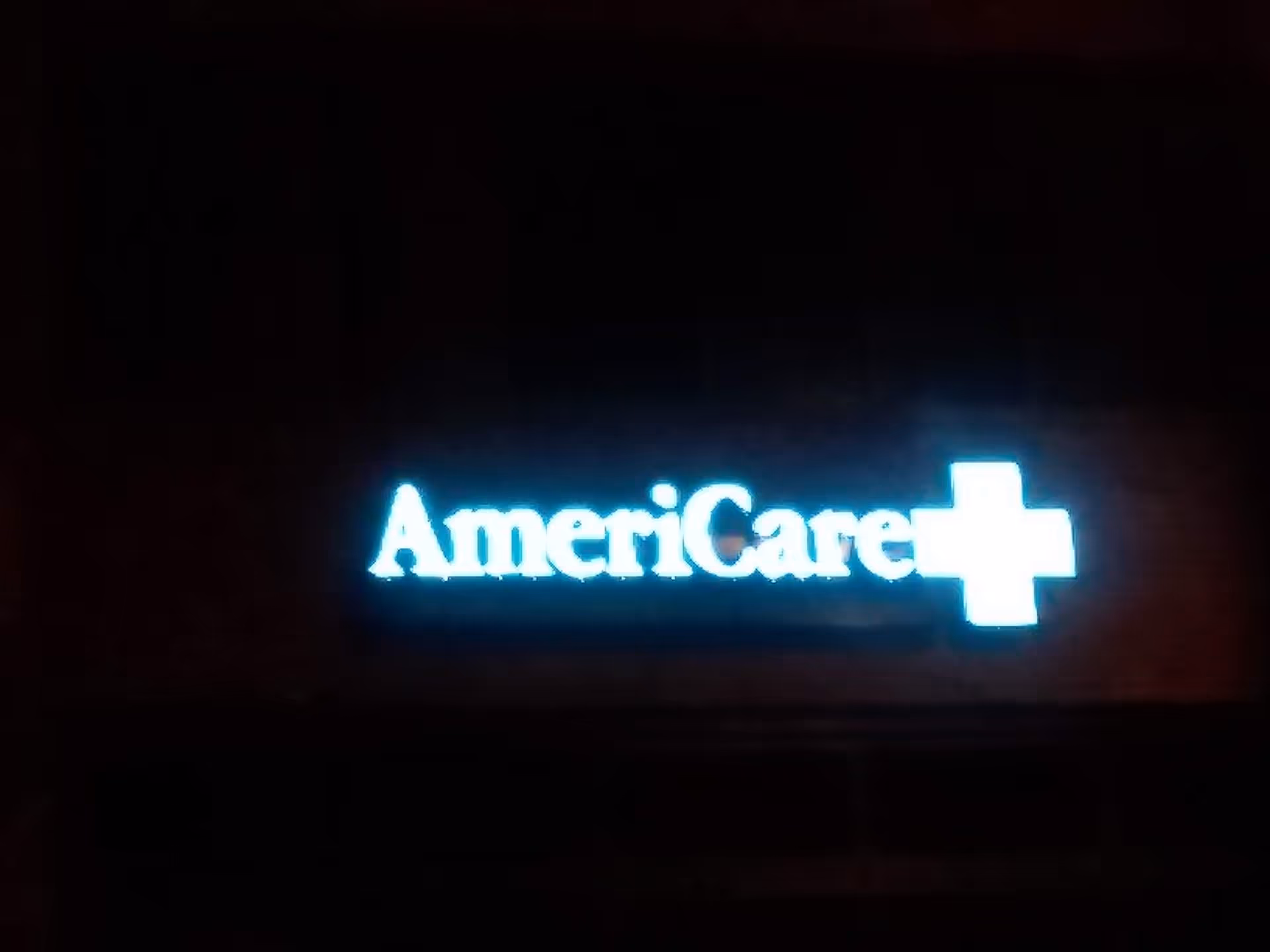 Blue illuminated AmeriCare sign with a cross on a dark building exterior