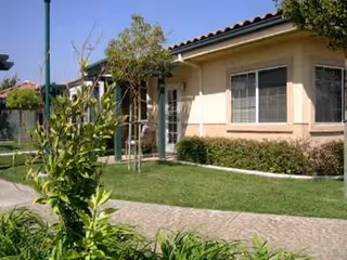 Single-story beige stucco building with windows, a tiled roof, manicured lawn, walkway, and small trees in front.