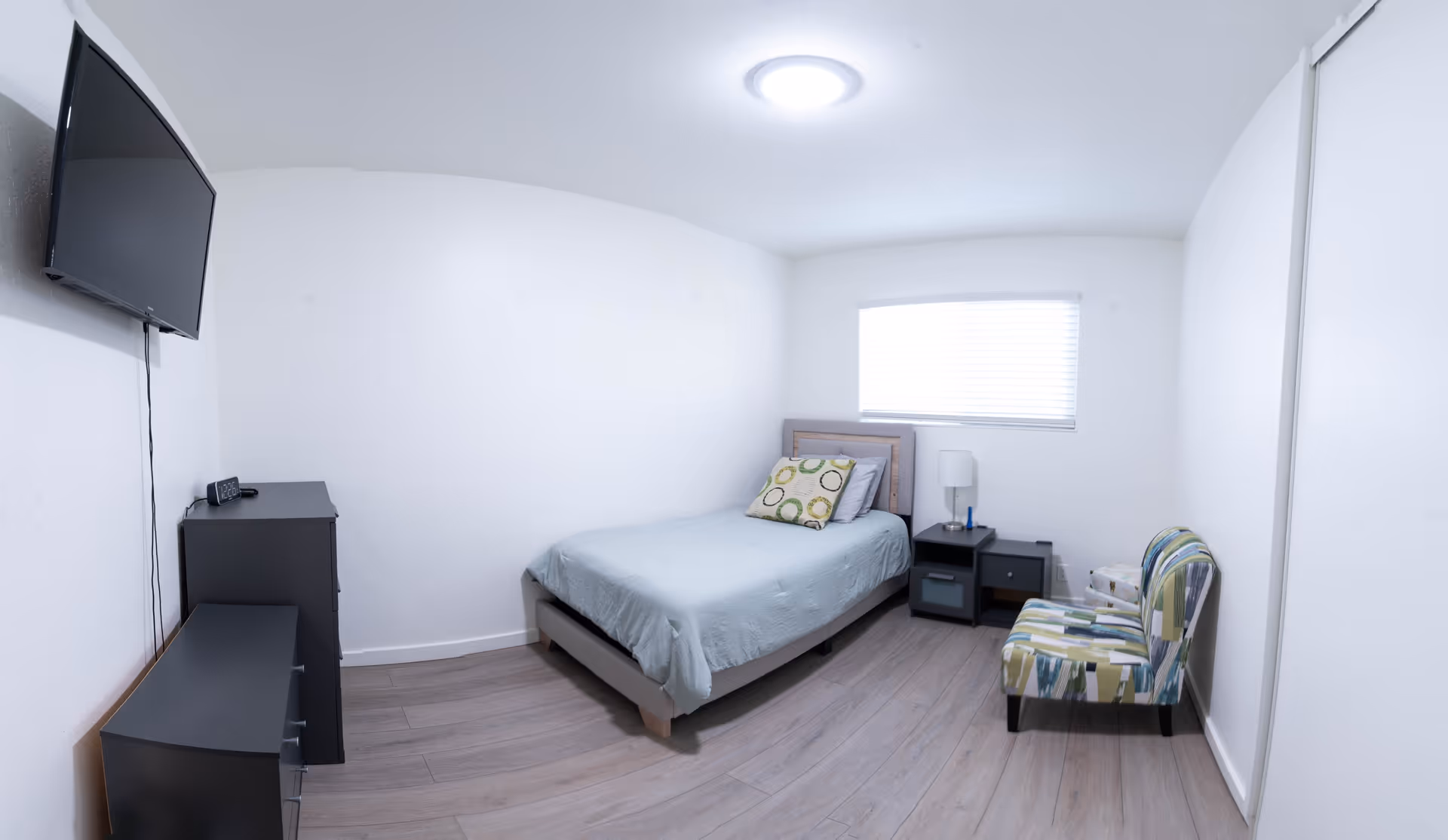 Single bedroom in a senior living facility with bed, dresser, nightstand, chair, and TV.