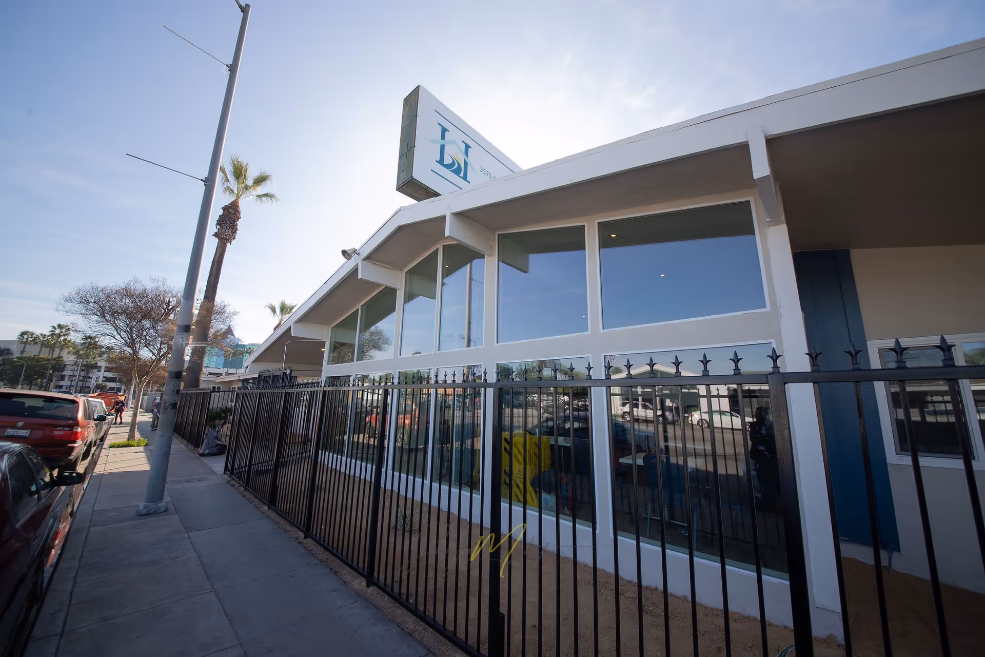 Exterior view of a single-story building with large windows and a black metal fence along the sidewalk. There are palm trees and parked cars along the street, and a sign on the roof with a logo and partial address visible.