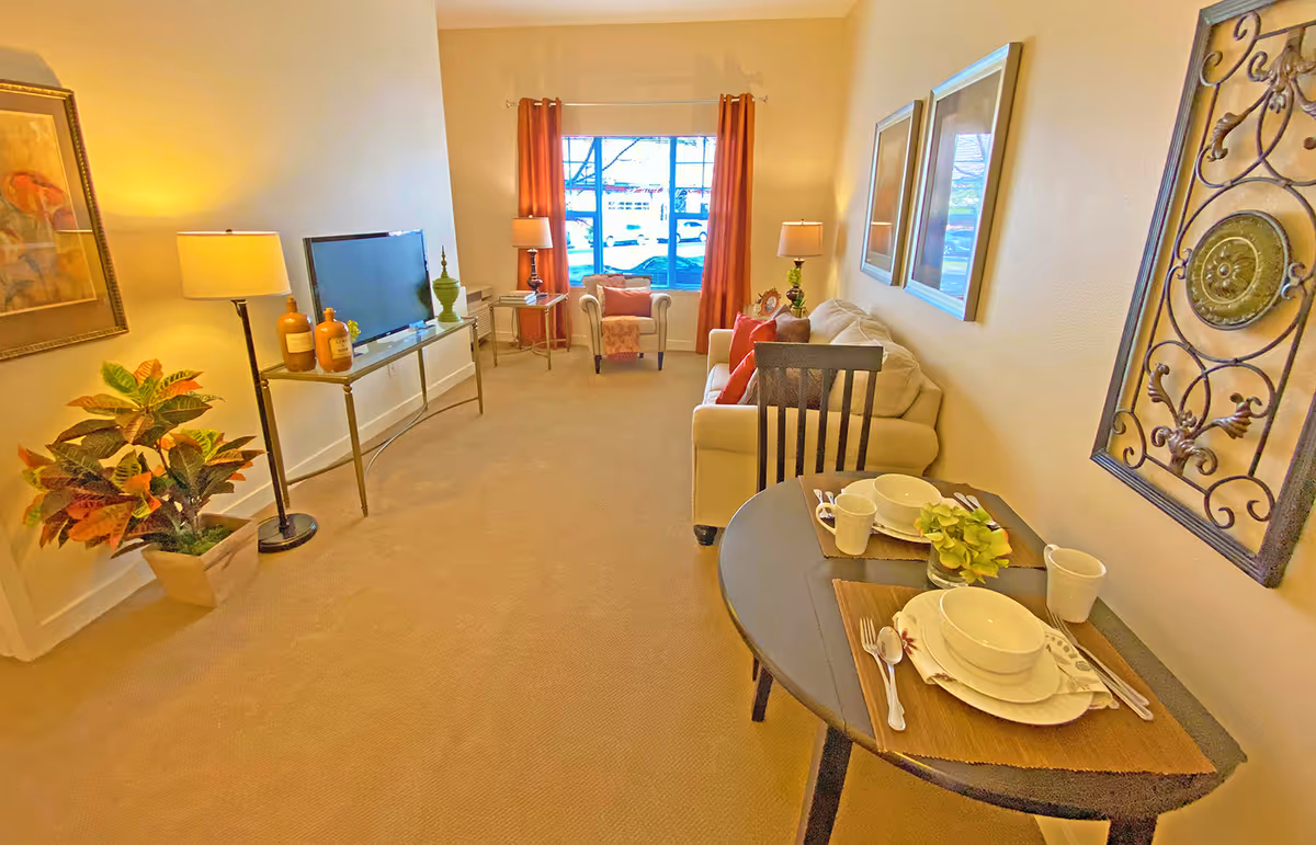 A cozy living room area with a beige sofa adorned with orange and beige cushions, a small round dining table set for two with white dishes and cups, a flat-screen TV on a slim console table, two floor lamps, a potted plant, and a large window with orange curtains letting in natural light.