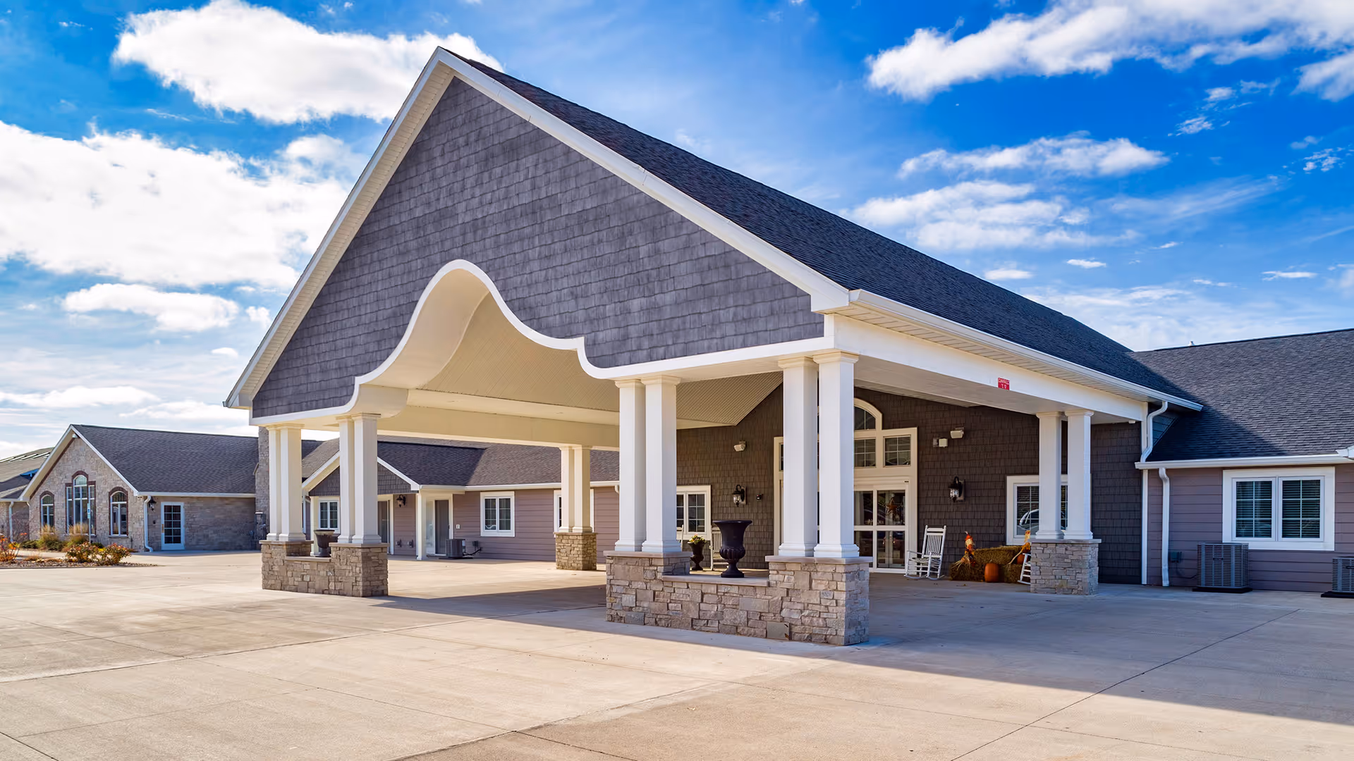 Front entrance of a single-story senior living facility with a large covered porte-cochere, white columns, stone accents, and a clear blue sky.
