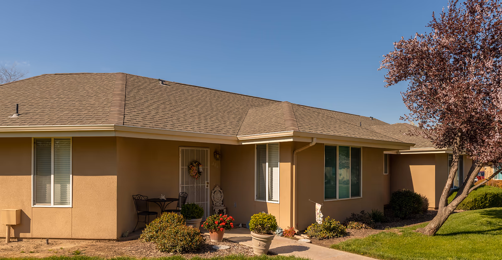 Single-story beige senior living building exterior showing the front entrance, potted plants, and a tree.