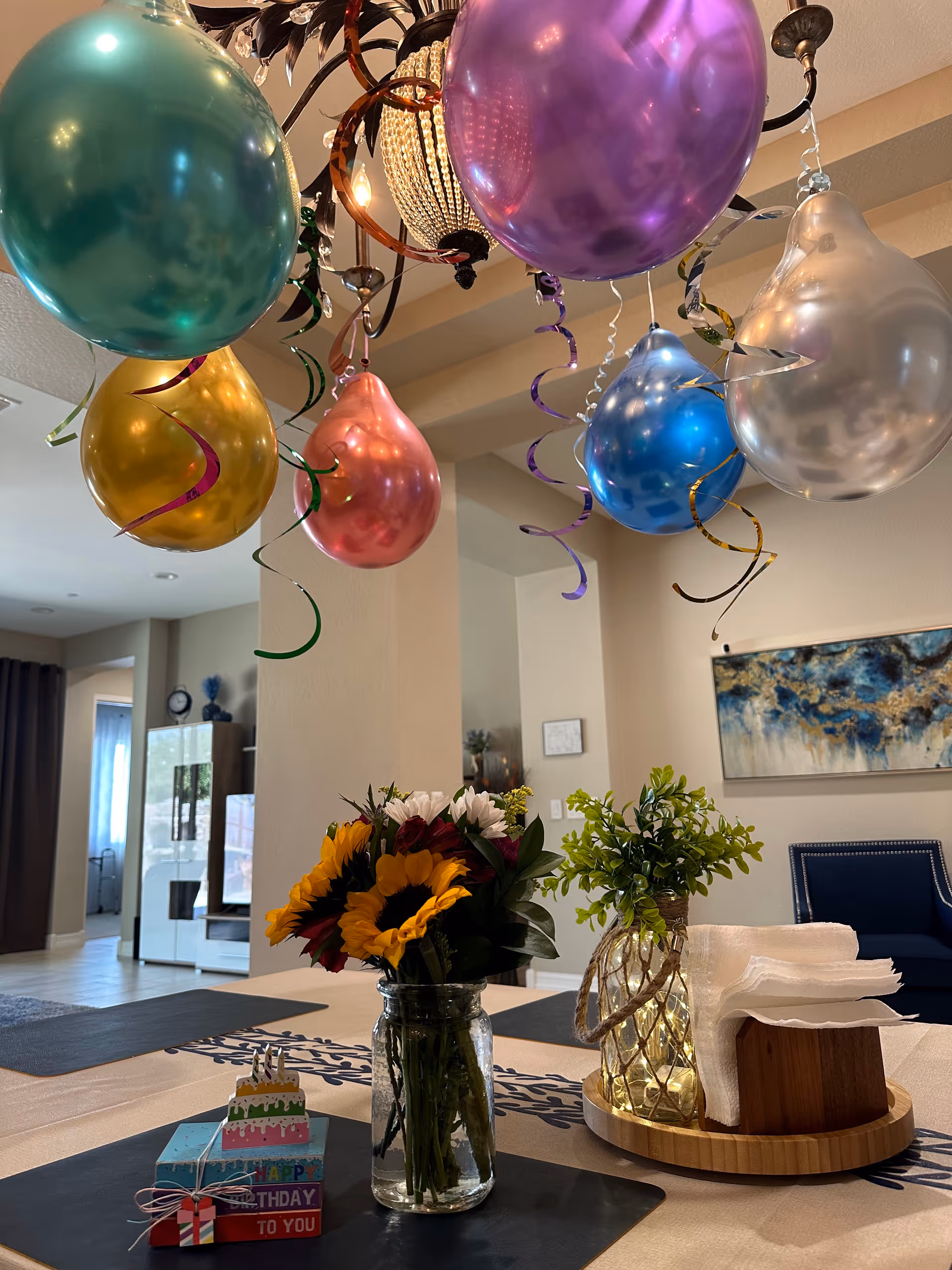 A decorated indoor space with colorful balloons hanging from the ceiling, a table with a vase of sunflowers and other flowers, a small stack of birthday-themed gift boxes, and a wooden tray holding napkins and a small plant. In the background, there is a blue armchair, a piece of abstract wall art, and a glimpse of other furniture and decor.