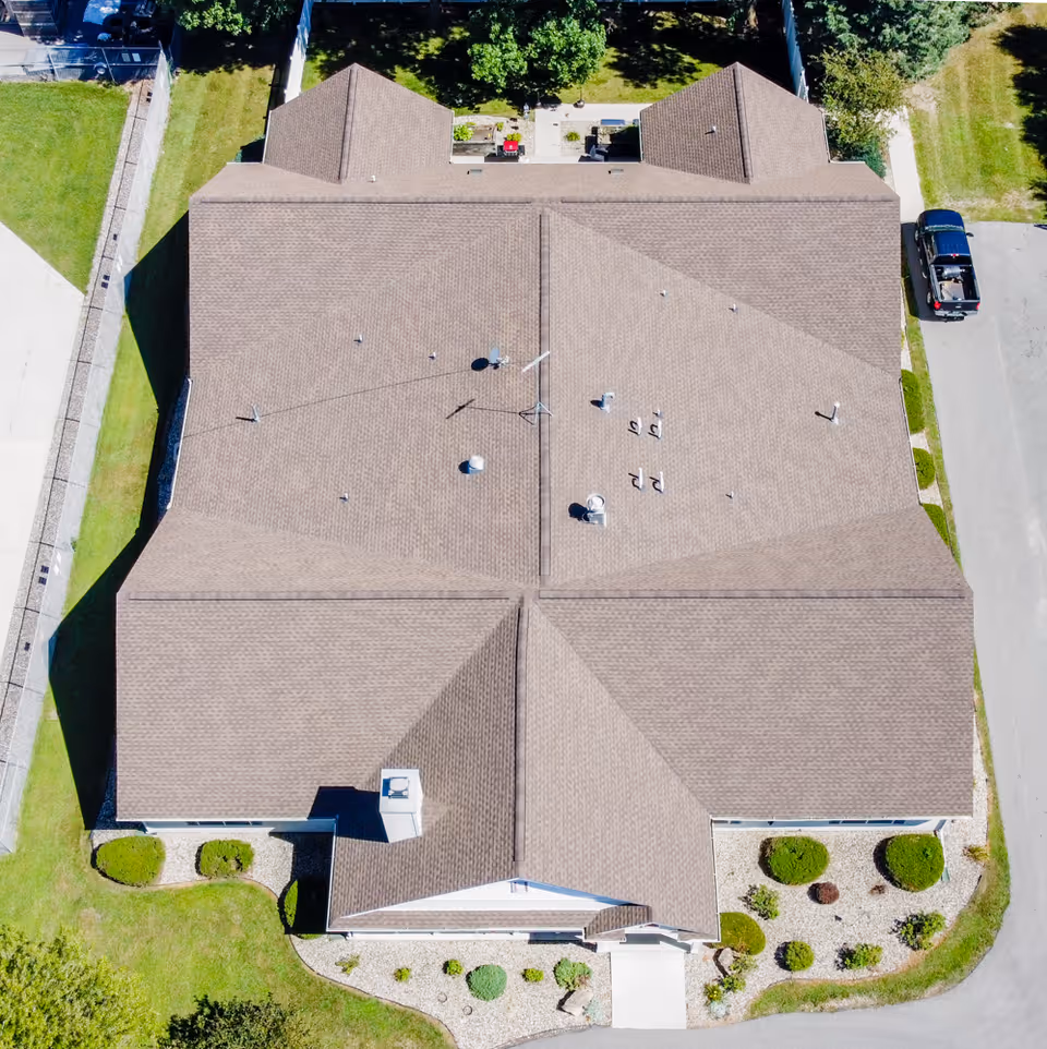 Aerial view of a large building with a brown shingled roof, surrounded by green lawns, bushes, and a paved road with a parked black pickup truck.