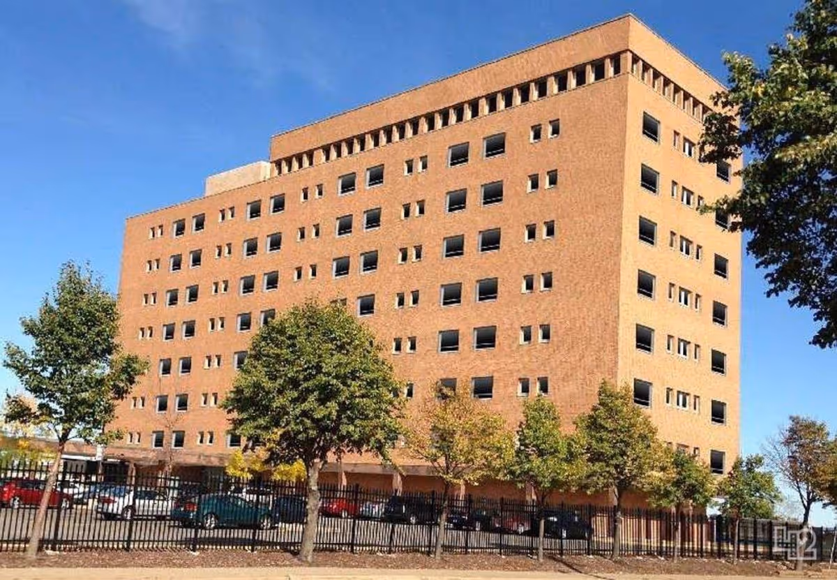 A large brick multi-story building with many windows, fenced parking, and trees under a blue sky.