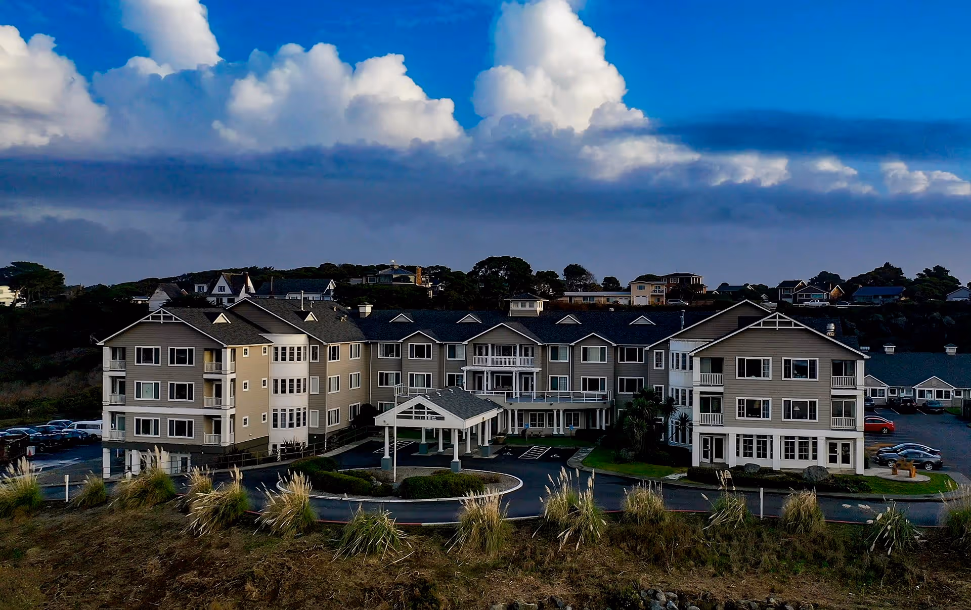 Exterior view of a large senior living community building with multiple floors, balconies, and a covered entrance. The building is surrounded by a parking lot and landscaped areas with grass and shrubs. The sky above is partly cloudy with large white clouds.