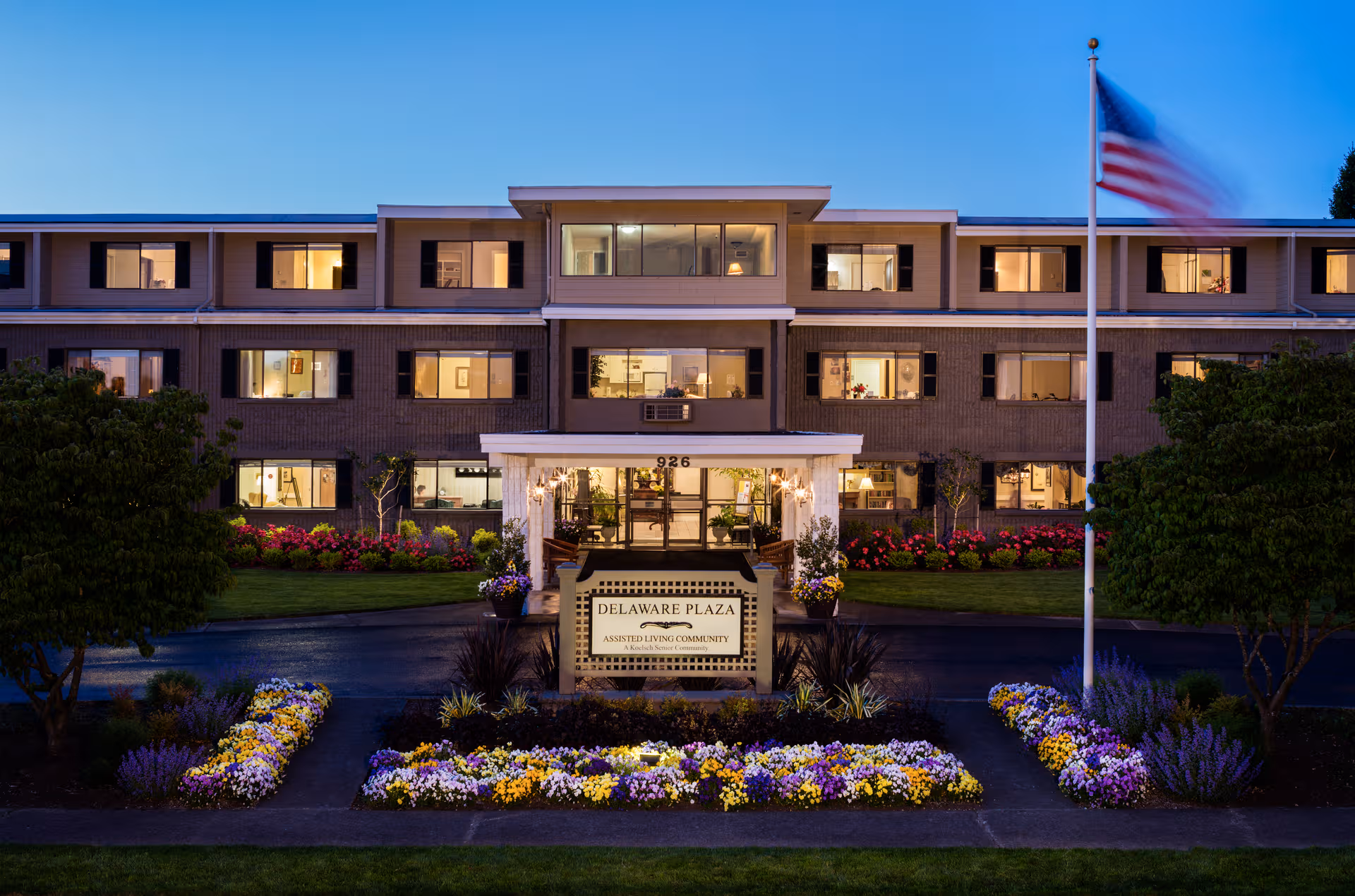 Exterior view of Delaware Plaza Assisted Living Community building with landscaped front and flag.