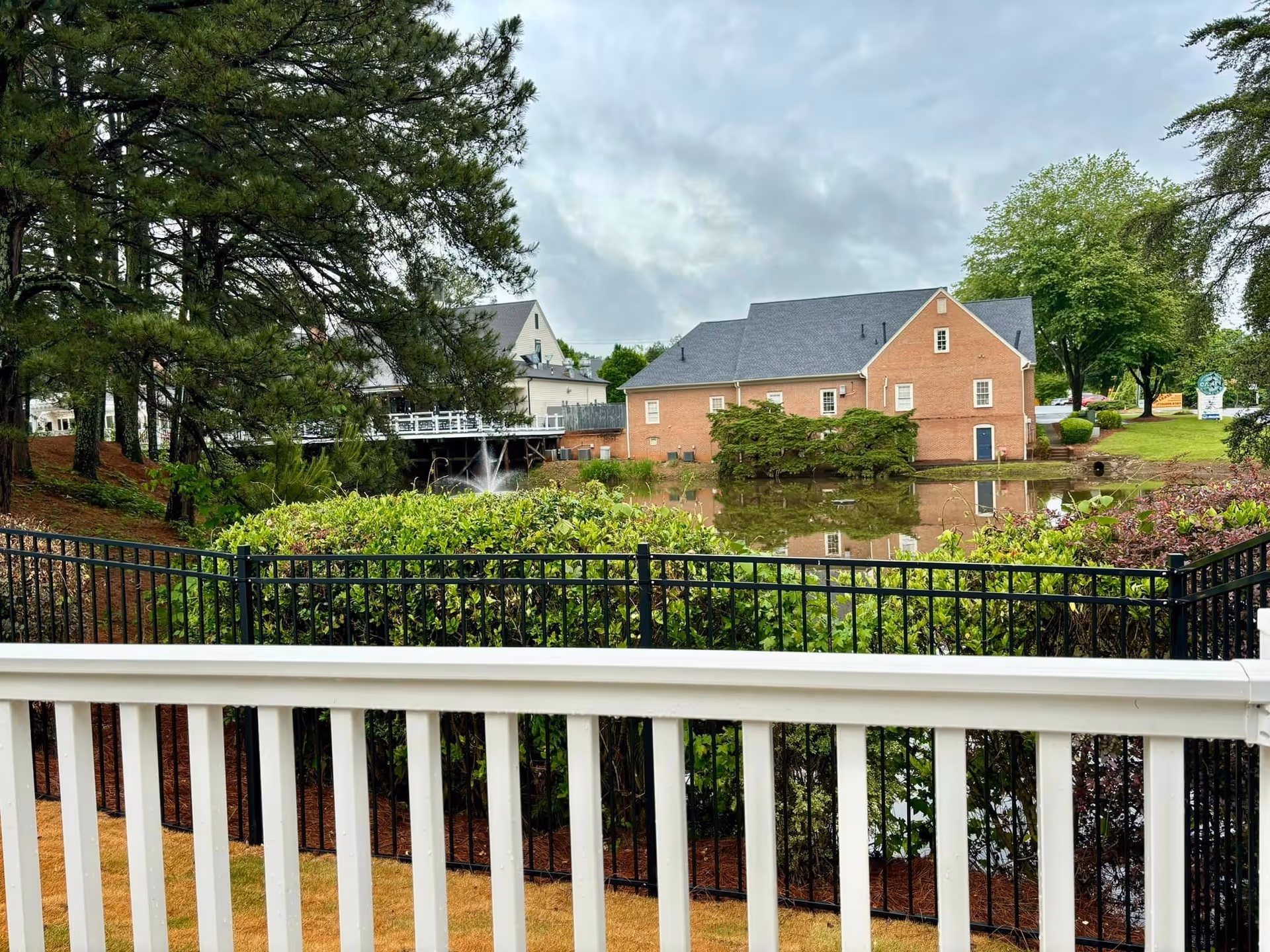 Brick senior living building across a pond, with trees, fence, and white railing in foreground.