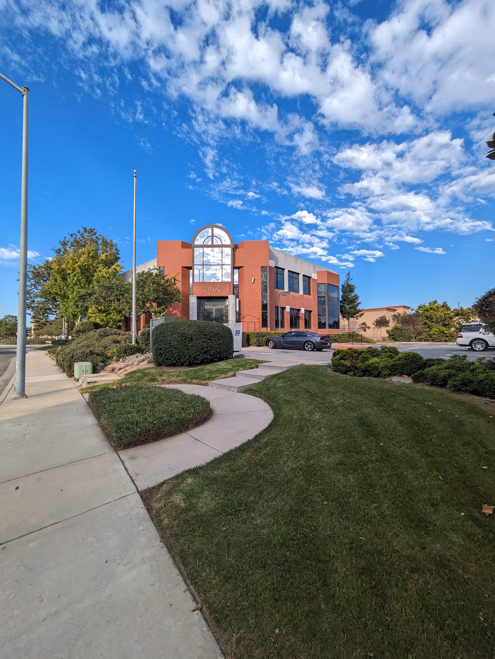 Red brick senior living building with curved glass entry, landscaped front, and blue sky.