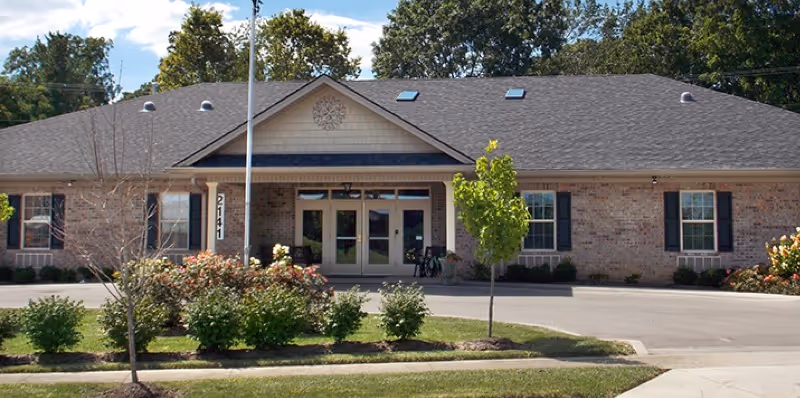 Front exterior of a single-story brick assisted living building with a central entrance, landscaping, and a driveway.