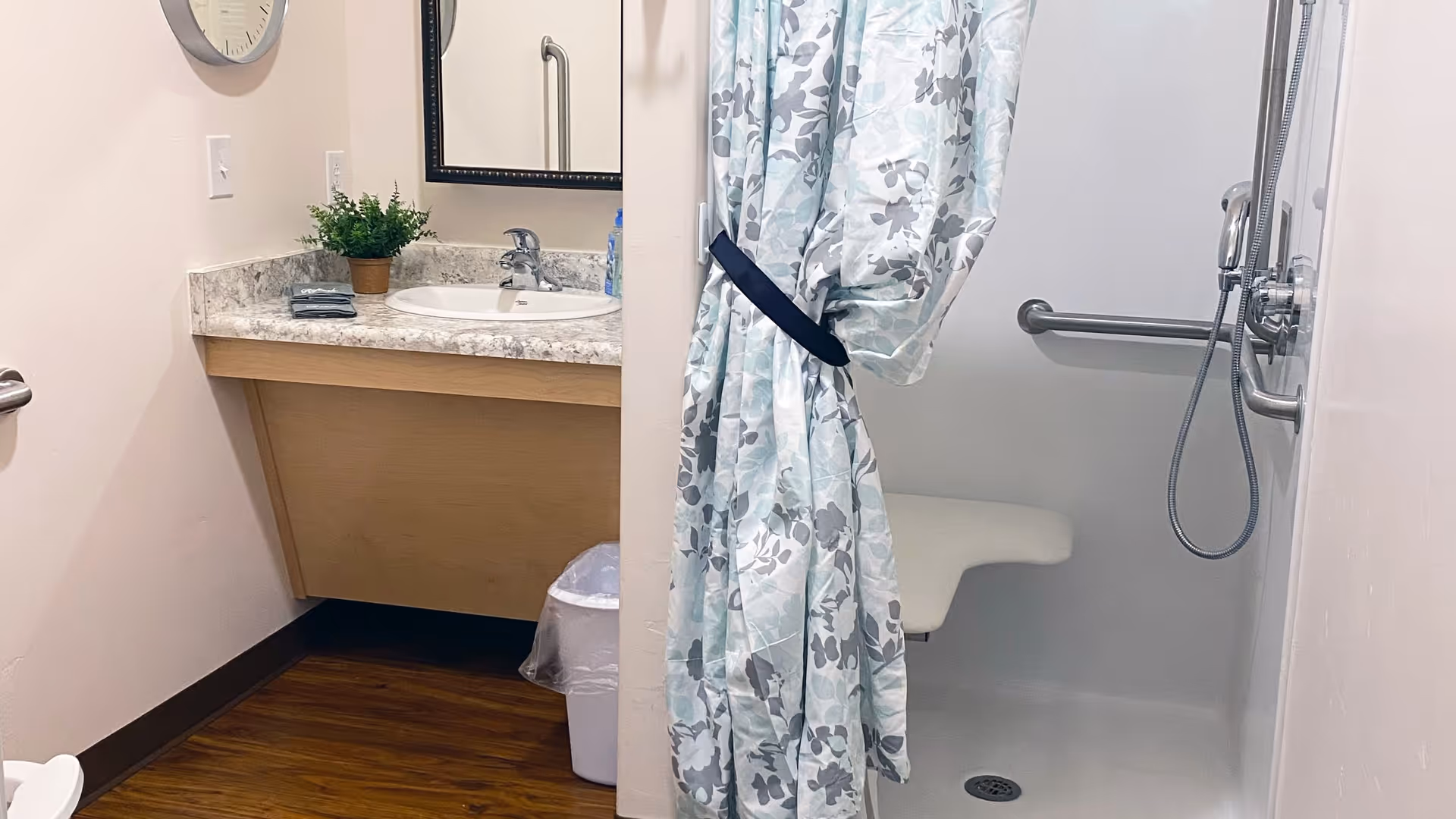 Senior living facility bathroom with sink, mirror, shower, and floral curtain.