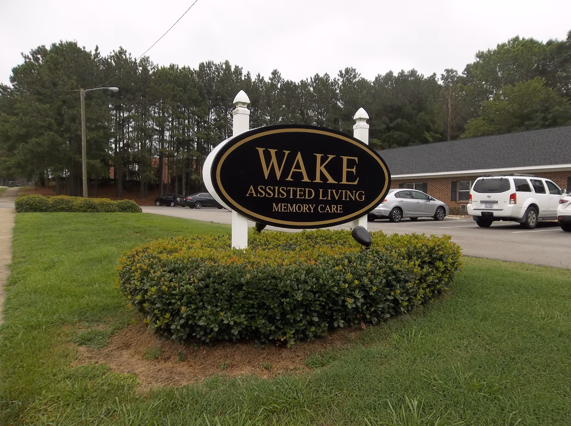Outdoor view of a sign for Wake Assisted Living Memory Care surrounded by trimmed bushes and grass, with a parking lot and several cars in the background along with trees and a building.