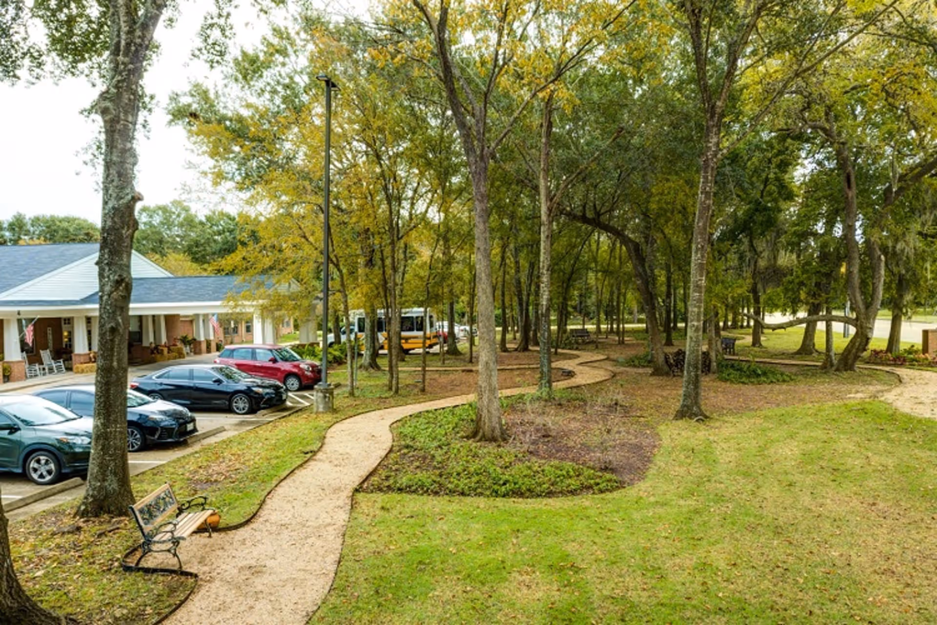 A winding gravel pathway through a wooded garden area with trees and green grass. There are benches along the path and a parking lot with several cars and a building with a covered porch in the background.