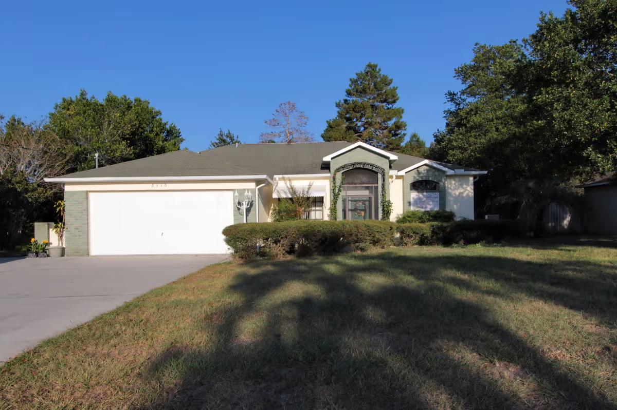 Single-story house-like building with an attached two-car garage, trimmed hedges and a front lawn under a clear blue sky.