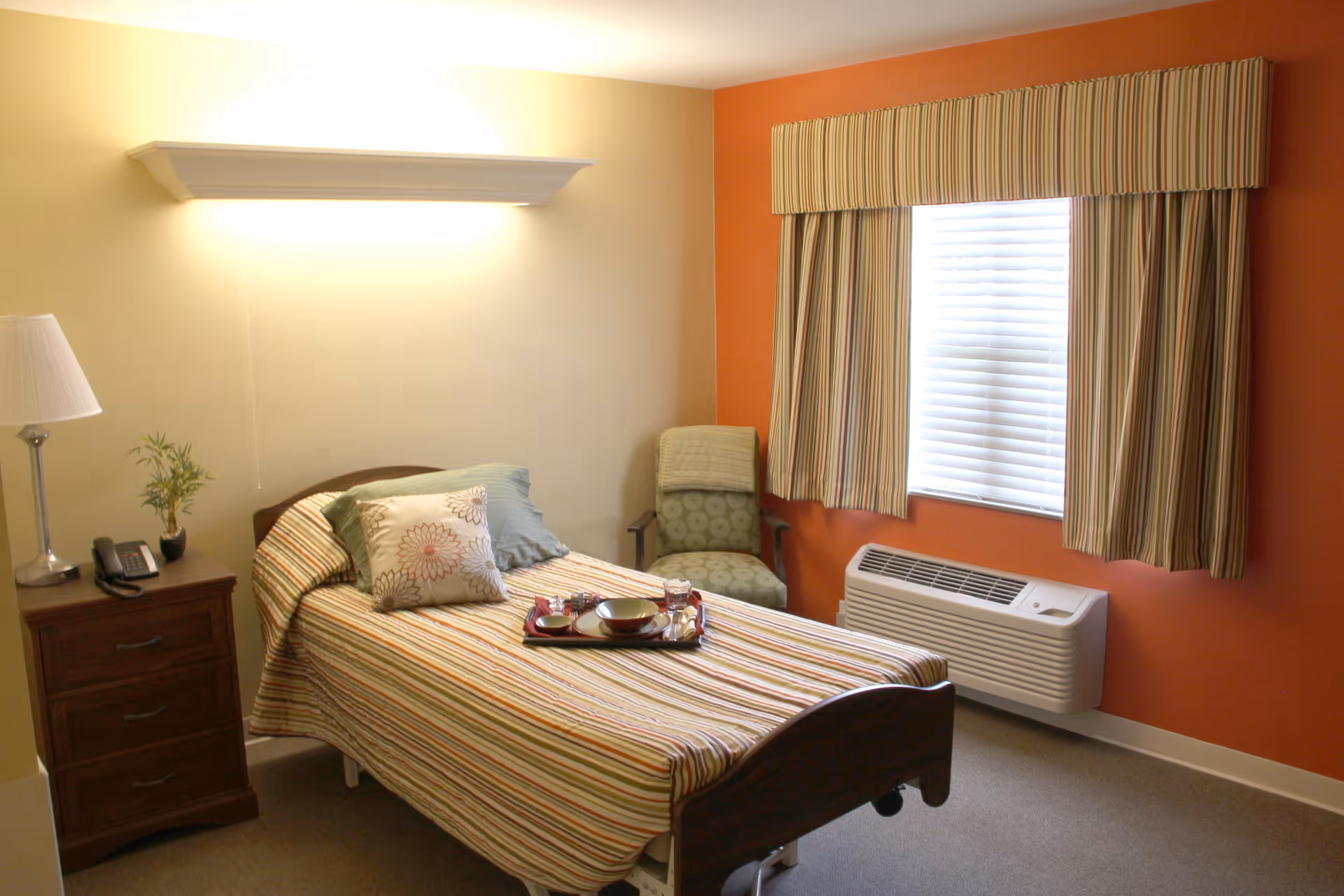 A cozy bedroom with a single bed covered in a striped bedspread and decorative pillows. A wooden nightstand with a lamp, phone, and small plant is beside the bed. There is a green cushioned chair next to a window with striped curtains, and a wall-mounted air conditioning unit below the window. The walls are painted in warm tones of beige and orange.