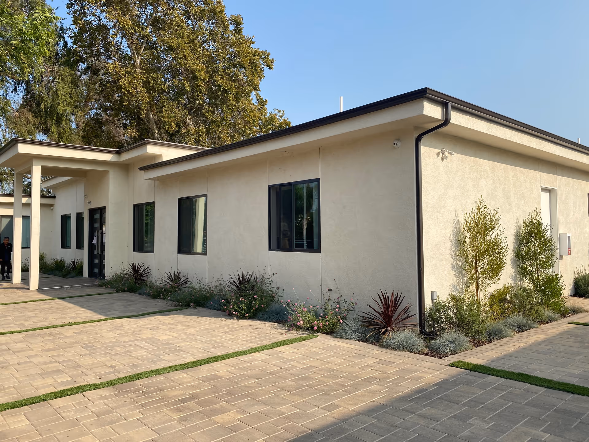 Exterior view of a single-story building with beige walls and several windows, surrounded by neatly landscaped plants and trees under a clear blue sky. The paved walkway and driveway lead to the entrance, where a person is visible near the door.